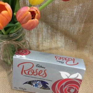 A 225g box of Cadburys Roses chocolates on a hessian background with a jar of tulips shown behind.